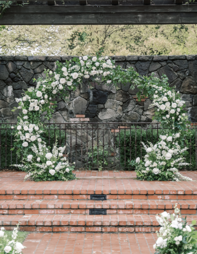 Wedding ceremony setup with a circular floral arch of white roses and greenery, brick steps, a stone wall, and rows of white chairs facing the arch.